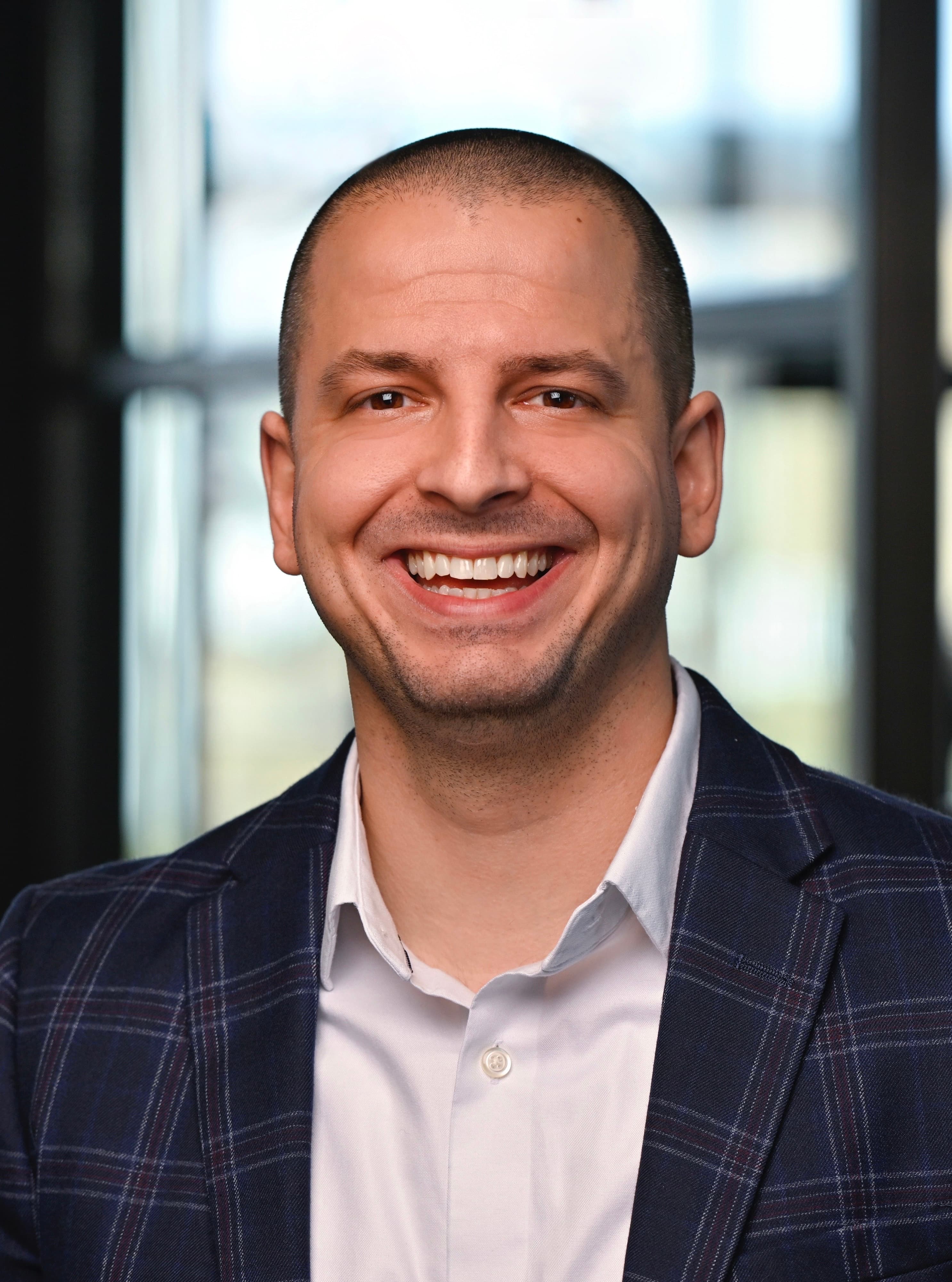 Smiling man with a buzzed haircut wearing a blue plaid blazer over a white shirt.